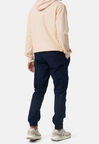 Marine cargobroek met elastische boorden, voorzien van zijzakken en een achterzak, gecombineerd met een lichtbeige hoodie en beige sneakers.