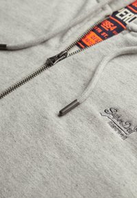 Grå zip-up hoodie lavet af blødt stof, med et kontrasterende indvendigt mærke med branding, en metal lynlås og en snor med metalspidser.