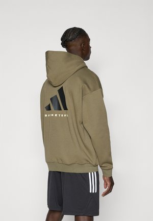 Mand med flettet hår iført en olivengrøn hættetrøje med et sort basketball-logo og sorte shorts med hvide striber, set bagfra.