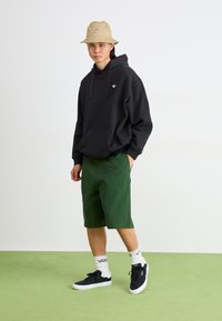 Μαύρο φούτερ με κουκούλα και καγκουρό τσέπη, πράσινες βερμούδες, μαύρα αθλητικά παπούτσια, λευκές κάλτσες και μπεζ καπέλο bucket με λογότυπο. Απλό φόντο.