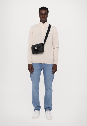 Helles beiger Zip-Kragen-Pullover, hellblaue Jeans, weiße Sneaker und eine kleine schwarze Umhängetasche mit silbernem Schnallen-Detail.