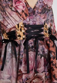 Robe en tissu à motifs floraux, mettant en avant une taille cintrée beige et noire avec des boutons dorés et des rubans noirs pour les ajustements.