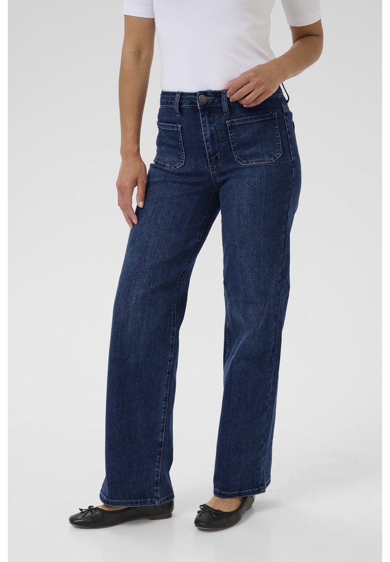 Tumesinised laia lõikega teksad kõrge vöökohaga, viie taskuga ja nähtavate õmblustega; kombineeritud mustade madalate kingade ja valge topiga.