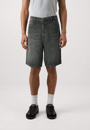 Mand iført knælange sorte denimshorts, en lysegrå t-shirt, hvide sokker og sorte sneakers står op ad en ensfarvet baggrund.