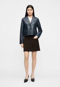 Γυναίκα στέκεται φορώντας ένα σκούρο μπλε δερμάτινο μπουφάν, σκούρα καφέ φούστα με μπροστινές τσέπες, λευκό τοπ και μαύρες μυτερές γόβες μπροστά σε απλό φόντο.