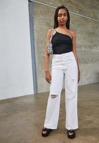 Top preto assimétrico, jeans brancos de perna larga com rasgos, e sandálias plataforma pretas. Bolsa de ombro com padrão de zebra adiciona contraste. Fundo simples.