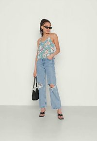 Top de alças finas em teal com estampado floral, combinado com jeans largo de perna larga em azul claro e rasgado. A modelo segura uma bolsa preta e usa sandálias pretas.