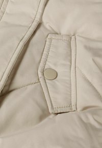 Beige vadderad tyg med en slät yta, som har en knappstängning och diagonala sömmönster, vilket framhäver värme och komfort.