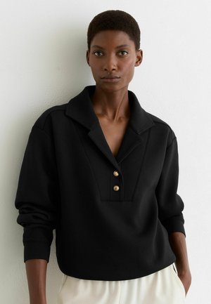 Donna con capelli corti che indossa una maglia nera a maniche lunghe con un ampio colletto e due bottoni dorati, in piedi contro una parete bianca.