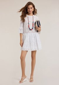 Rochie albă din dantelă cu mâneci trei sferturi, fustă evazată și modele brodate; asortată cu un colier colorat din mărgele și o poșetă.