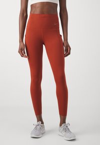 Wysokie legginsy dopasowane w kolorze rdzy, wykonane z elastycznego materiału, posiadające boczne kieszenie i logo Nike na pasku.