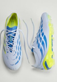 Chaussures de football blanches et bleues avec des accents jaunes et des crampons translucides, une chaussure lacée et l'autre avec des lacets défaits à côté.