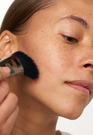 Un pinceau à maquillage avec une tête en poils noirs applique du produit sur la joue d'une personne à la peau brun clair et aux taches de rousseur visibles.