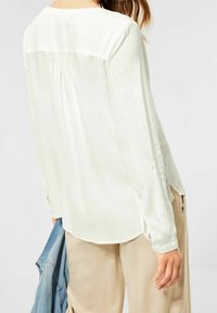 Blusa a maniche lunghe color crema con scollatura rotonda, dettaglio plissettato sulla schiena e polsini con bottoni, abbinata a pantaloni beige e a una giacca di denim chiaro.