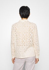 Camicia color beige chiaro con motivi floreali traforati, maniche lunghe e colletto. Tessuto morbido con una vestibilità rilassata. Indossata sopra pantaloni di colore chiaro.