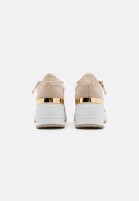 Ett par beige plattformssneakers med metalliska guldackenter och vita chunky sulor, visade från baksidan på en vit bakgrund.