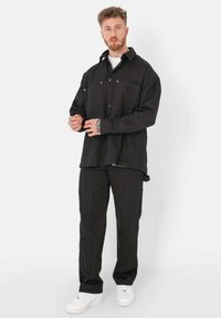 Chemise noire oversize avec fermeture à boutons, dotée de deux poches avant et d'un col. Pantalons noirs avec poches latérales et baskets blanches.