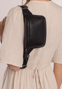 Borsa a tracolla in pelle nera con scomparto con zip e tracolla regolabile, indossata sulla schiena di una persona, caratterizzata da una texture liscia.