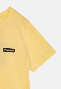 Kollane puuvillane t-särk lühikeste varrukatega, mille esiküljel on must ristkülikukujuline silt "VINGINO" tekstiga. Sile tekstuur, vabaltaja disain.