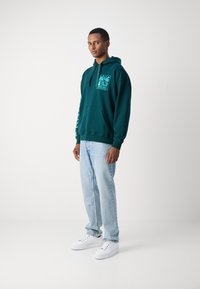 Felpa verde acqua con tasca frontale e stampa grafica, abbinata a jeans azzurri chiari e sneakers bianche; vestibilità rilassata e morbida texture.