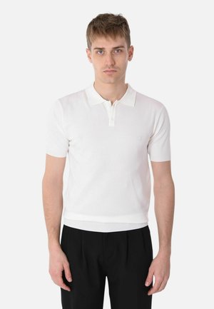 Giovane uomo con capelli corti in piedi di fronte, indossa una polo bianca a maniche corte e pantaloni neri su uno sfondo semplice.