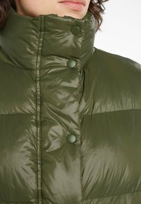 Grüne gepolsterte Jacke mit hohem Kragen, glänzender Textur und Druckknopfverschluss. Der Stoff wirkt leicht und hat ein gestepptes Design.