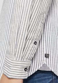 Camisa de botones a rayas con rayas verticales en gris y blanco, con un detalle de botón negro en el puño y una etiqueta en el dobladillo.