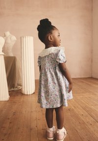 Kleid mit Blumenmuster, kurzen Puffärmeln, Spitzenkragen und plissiertem Rücken. Kombiniert mit rosa Schuhen und weißen Rüschenstrümpfen auf Holzboden.