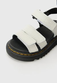 Hvitt lær sandaler med tre justerbare borrelåsremmer, svart såle med teksturert overflate, og gult sømdetalj langs kanten.