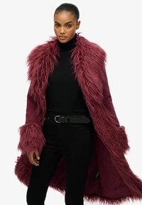 Burgundy nepvacht coat met een gestructureerd ontwerp, met een brede kraag en manchetten, gedragen over een zwarte coltrui en zwarte broek.