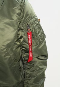Olive green flight jacket in shiny nylon fabric, featuring a zippered pocket and a red strap labeled "REMOVE BEFORE FLIGHT."