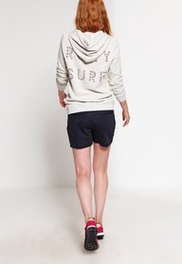 Žena s dlouhými červenými vlasy, která odchází, má na sobě světlou mikinu s textem, tmavé kraťasy a růžové tenisky na jednobarevném bílém pozadí.