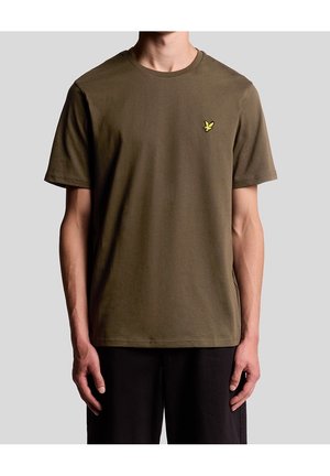 Mężczyzna ubrany w oliwkowozieloną koszulkę z krótkim rękawem z małym żółtym logo ptaka na lewej piersi oraz czarne spodnie na jednolitym tle.