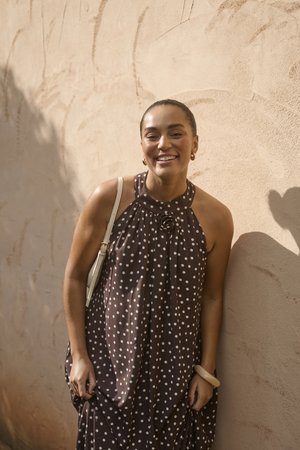 Femme souriante en robe marron sans manches à pois, portant un sac à bandoulière blanc et un bracelet, debout contre un mur beige texturé au soleil.