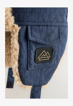 Gorro de tela acolchada azul con forro de piel sintética color beige y un parche con gráficos de montañas; incluye un bolsillo lateral para almacenamiento.