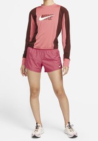 Nike long-sleeve top in pink and brown with logo; paired with pink patterned shorts. Model wears light sneakers with black details.