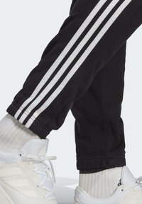 Svarta träningsbyxor med tre vita ränder nedanför sidan, åtsittande muddar, bärs med vita sneakers och ribbade strumpor.