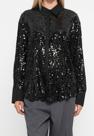 Kobieta w czarnej, długorękawowej cekinowej koszuli na guziki z kołnierzykiem i szarych spodniach, stojąca na jednolitym tle.