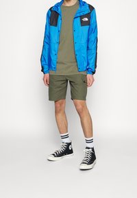 Blå och svart vindjacka, olivgrön t-shirt, khakishorts, svarta högklackade sneakers med vita sulor, randiga strumpor.