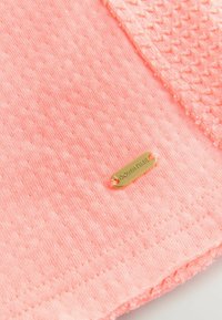 Tissu en corail doux avec un motif texturé, doté d'une étiquette dorée avec "DOWNTIME" et de délicates coutures le long des bords.