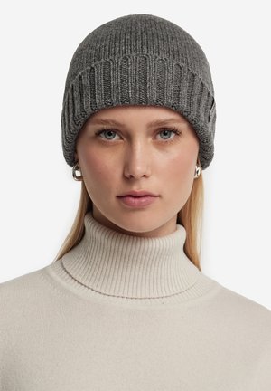 Grey knitted beanie with ribbed texture, featuring a rolled brim and a small logo accent. Worn with a light beige turtleneck sweater.