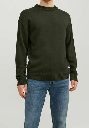 Personne portant un pull tricoté vert foncé et un jean bleu, debout avec les bras détendus sur un fond clair uni.