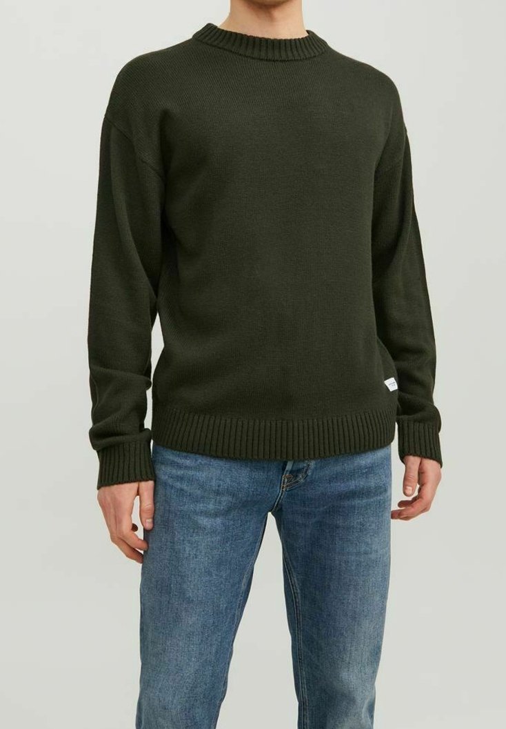 Personne portant un pull tricoté vert foncé et un jean bleu, debout avec les bras détendus sur un fond clair uni.