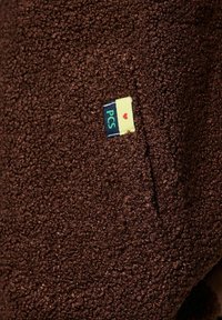 Brunt texturerat tyg med en mjuk, lockig yta. Har en liten etikett med "PCS" och ett hjärta på en gul och blå bakgrund.