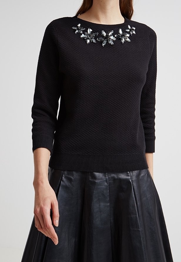 Femme portant un pull noir texturé à manches longues avec un col floral orné de bijoux, associé à une jupe en cuir plissée noire.