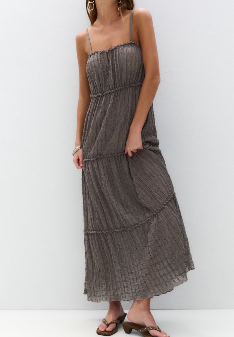 Femme portant une robe longue grise texturée sans manches avec de fines bretelles et des sandales marron à talons, debout contre un mur blanc uni.