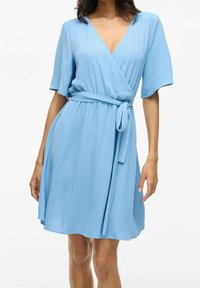 Woman wearing light blue wrap dress with short sleeves and fabric belt tied at the waist, standing against a plain background.