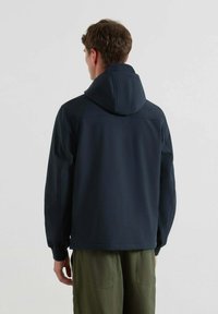 Uomo che indossa un giubbotto con cappuccio blu scuro e pantaloni verde oliva, in piedi di spalle contro uno sfondo chiaro e uniforme.