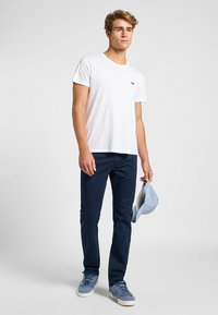 T-shirt bianca con un piccolo logo nero, pantaloni blu scuro, sneaker blu e un cappellino azzurro tenuto in mano. Design semplice, vestibilità informale.