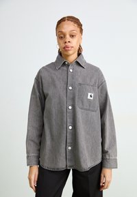 Camicetta in denim grigio con colletto, grande tasca frontale e bottoni bianchi. Tessuto morbido con una vestibilità rilassata e dettagli di cuciture a contrasto.
